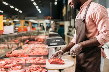 cropped image of african american male shop assistant in apron cutting raw meat in grocery store - obrazy, fototapety, plakaty