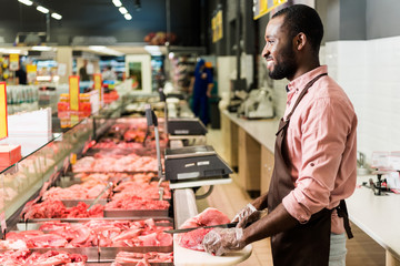 side view of smilng african american male butcher in apron cutting raw meat in grocery store