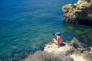 Back side view of person sitting holding tablet computer on ocean sunny natural background. Traveler having fun on international travelling lifestyle, technology social communication, amazing nature.