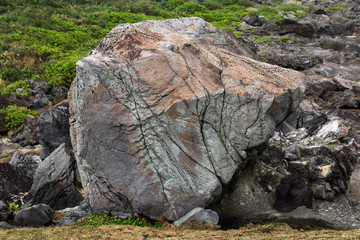 Image of a single large rock boulder, peach, salmon and gray/grey in color with multiple cracks in the side. Boulder is sitting on some brown grass, with green grass and small rocks in background.