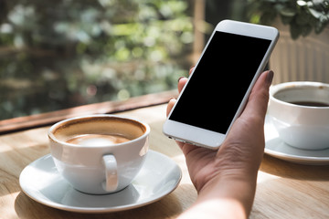 Mockup image of hand holding white mobile phone with blank black desktop screen with coffee cup on wooden table in cafe