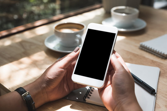 Mockup image of hands holding white mobile phone with blank black desktop screen with notebook and coffee cup on wooden table in cafe