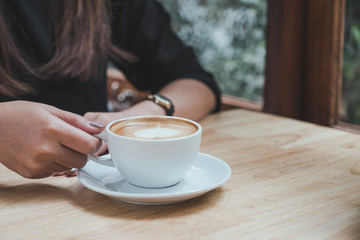 Closeup image of a woman holding a white cup of hot coffee on wooden table in cafe