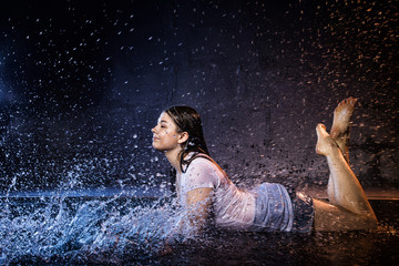 Girl with wet hair in the white shirt, water drops around and dark wall background illuminated by...
