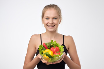 Smiling blonde young woman holding a pile of healthy fresh vegetables standing on white background. Concept of healthy diet eating