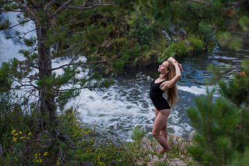 Beautiful young woman standing near the waterfall. A girl in a black swimsuit standing next to a waterfall.