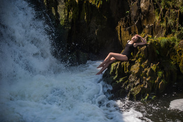 Young blond woman in a black swimwear sitting by the waterfall.  Young woman enjoying nature