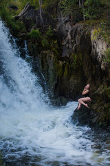 Young blond woman in a black swimwear sitting by the waterfall.  Young woman enjoying nature