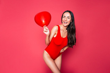 Happy Beautiful young smiling woman with long curly hair in red swimsuit posing over pink background with red balloon in hand 