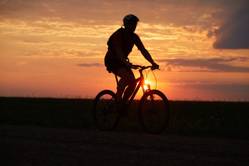 Fototapeta na wymiar Silhouette of a bicyclist traveling on a sunset background on the road to the field
