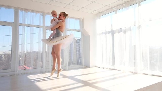 Happy mother in ballet outfit standing on tiptoe and holding on hands small daughter. Young ballerina is walking on tiptoe and raising her daughter up high