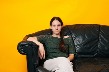 Young serios girl sitting on a black couch and looking at the camera on yellow background.