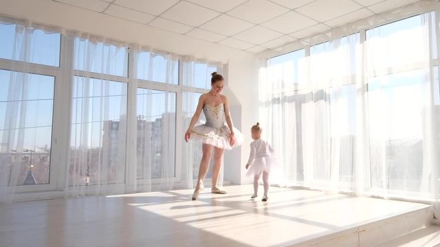 Young elegant ballerina teaches her little daughter dancing in studio. Happy ballerina with her child practice a ballet moves. Mother and daughter dancing ballet