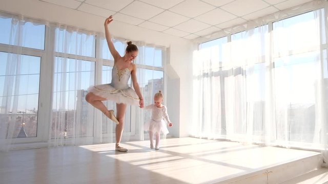 Young impressive ballerina teaches her little daughter dancing in studio. Happy ballerina with her child practice a ballet moves. Mother and daughter dancing ballet