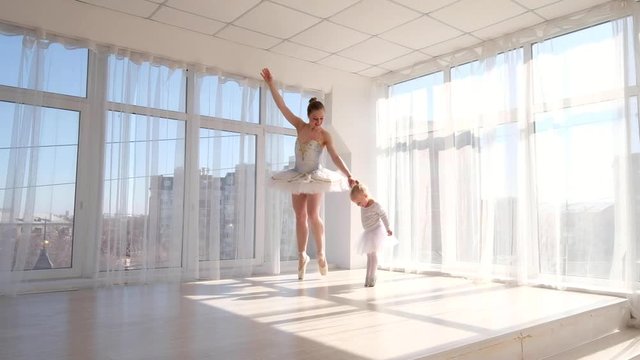 Young charming ballerina teaches her little daughter dancing in studio. Happy ballerina with her child practice a ballet moves. Mother and daughter dancing ballet
