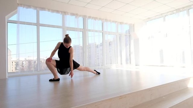 Attractive young ballerina in black swimsuit doing exercises for stretching in ballet school