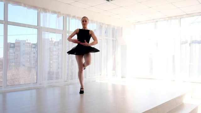Young attractive ballerina in black leotard practising ballet moves in white bright room
