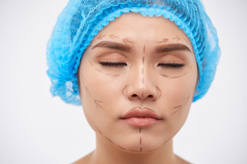 Young Asian woman prepared for decorative surgery standing with eyes closed and drawings on face