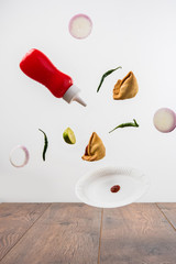 Tasty Samosa Snacks flying with ketchup bottle, green chilli, onion slices and plate over plain background