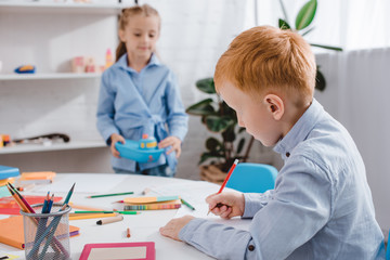 Fototapeta premium selective focus of red hair boy drawing picture while classmate standing near by at table