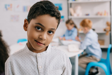 selective focus of cute african american boy looking at camera with classmates behind in classroom