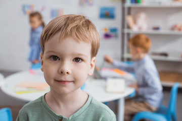 selective focus of cute little boy looking at camera with classmates behind in classroom