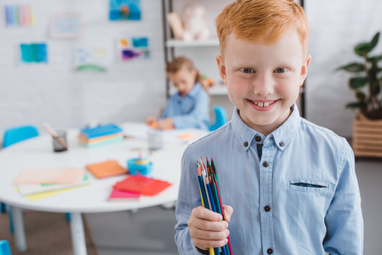selective focus of happy red hair boy with pencils and classmate at table in classroom