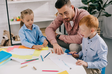 teacher in eyeglasses helping preschoolers with drawing at table in classroom