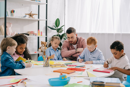 teacher and interracial preschoolers at table with paints and papers in classroom