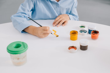 partial view of child drawing picture with paints and paint brush at table