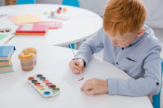 focused preschooler red hair boy drawing picture at table in classroom