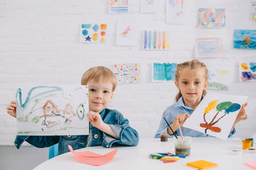 Fototapeta premium portrait of cute kids showing drawings in hands at table in classroom