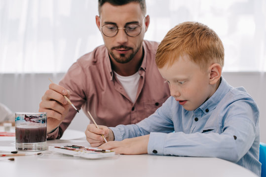 portrait of teacher helping red hair boy to draw picture at table in classroom