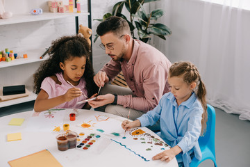 teacher in eyeglasses and multiracial children drawing pictures with paints at table in classroom