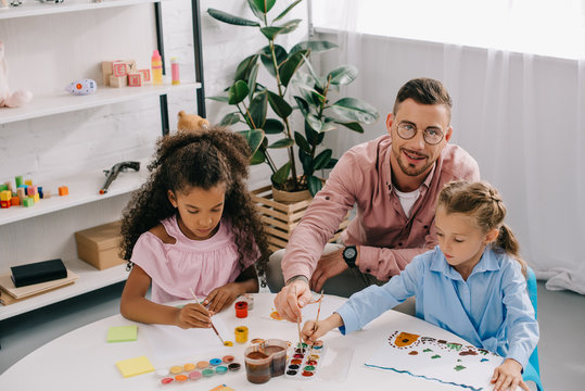 teacher in eyeglasses and multiracial children drawing pictures with paints at table in classroom