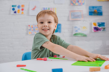 portrait of smiling boy sitting at table with colorful plasticine for sculpturing in room