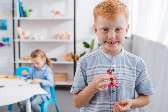 selective focus of smiling red hair boy with figure made of plasticine and classmate drawing picture at table in classroom