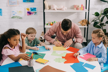 Fototapeta premium multiethnic preschoolers and teacher making paper applique at table in classroom