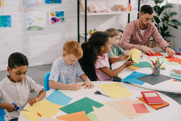 teacher and multicultural preschoolers cutting colorful papers with scissors in classroom