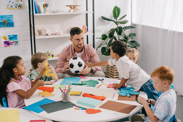 male teacher with soccer ball and multiracial preschoolers sitting at table with colorful papers in...