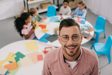 selective focus of smiling teacher in eyeglasses and interracial kids at table in classroom