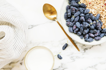 Fresh ripe honeysuckle berries and muesli in a ceramic bowl and cup of milk on a grey marble table....