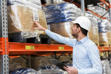 logistic business, shipment and people concept - businessman in helmet with clipboard checking goods at warehouse