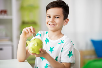 money, finances, childhood and people concept - smiling little boy putting coin into piggy bank at...