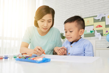 Young woman teacher and cute Asian boy drawing with crayons in art class in backlit