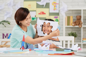 Asian teacher putting paper with flower cut-out to face of little girl in art class