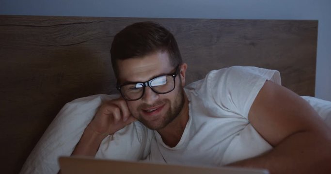 Close up of the smiled man using laptop computer and smiling while watching something in the evening in his bed.