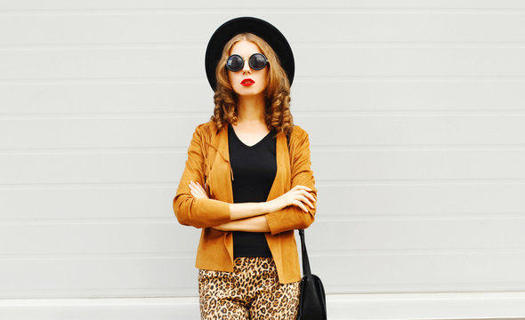 Elegant young woman wearing a retro elegant hat, sunglasses, brown jacket and black handbag clutch over grey background