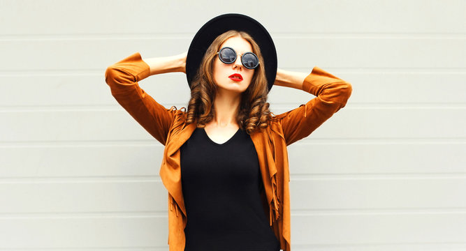 Elegant woman model wearing a black hat, sunglasses and jacket over urban grey background