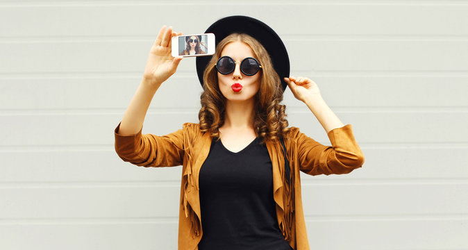 Cool girl model taking photo picture self-portrait on smartphone wearing retro elegant hat, sunglasses, brown jacket and handbag with curly hair over city grey background